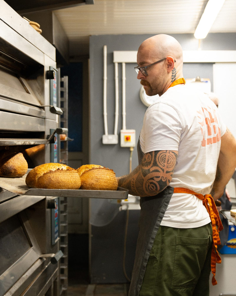 A Wildflour baker in an orange apron drawing a tray of sourdough loaves from the deck oven.
