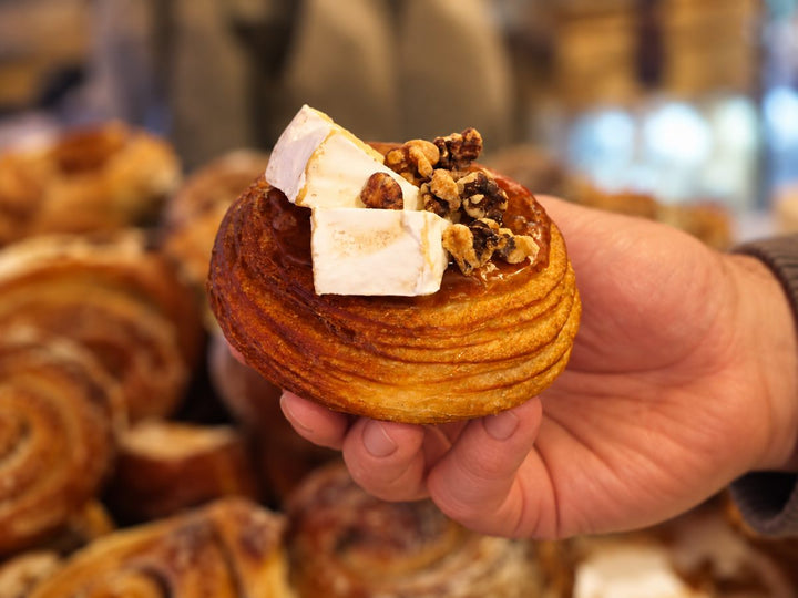 A round laminated pastry topped with wedges of brie and chopped walnuts, held up in front of trays of pastries at the counter.