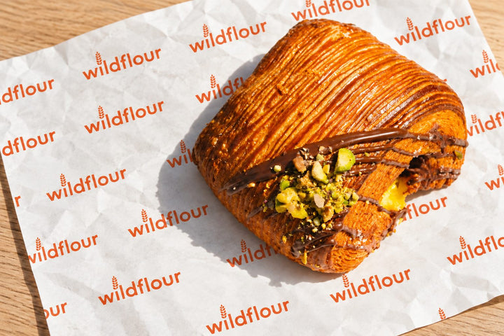 A pistachio and chocolate suisse on Wildflour branded paper, drizzled with dark chocolate and scattered with chopped pistachios.